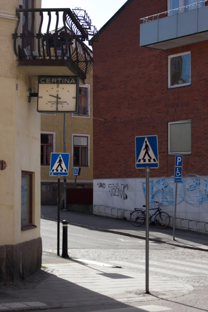 Gata i strängnäs med tre övergångsställesskyltar. Till vänster är en gammeldags klocka som hänger under en balkong